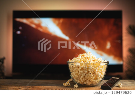 A glass bowl of popcorn and remote control in the background the TV works. Evening cozy watching a movie or TV series at home 129479620