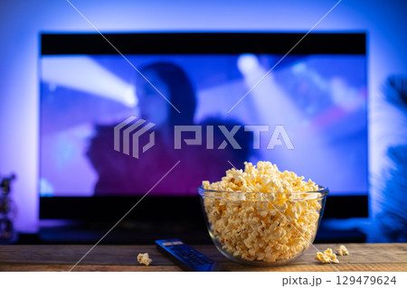 A glass bowl of popcorn and remote control in the background the TV works. Evening cozy watching a movie or TV series at home A glass bowl of popcorn and remote control in the background the TV works. Evening cozy watching a movie or TV series at home 129479624
