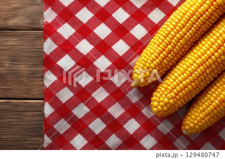 Three ears of fresh yellow corn lie on a red and white checkered tablecloth. The setting evokes a rustic kitchen atmosphere, ideal for summer meals and gatherings. 129480747