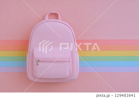 A pink backpack is centered against a pastel striped backdrop featuring soft rainbow colors. The backpack has a front pocket and a simple design, creating a casual aesthetic. 129481641