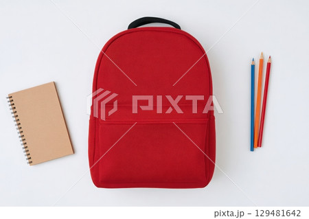 A bright red backpack is positioned next to a spiral notebook and colorful pencils on a clean white surface. This setup showcases essential school supplies in a simple layout. 129481642