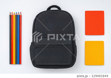 A black backpack sits in the center of a clean, white surface, flanked by rows of colored pencils in red, blue, yellow, and orange. 129481644
