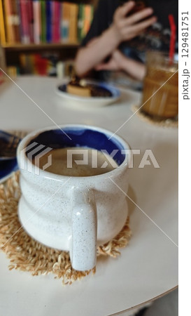 Cup of hot coffee in a ceramic mug on a white table in a cozy cafe with bookshelves in the background. Concept of relaxation, reading and coffee culture Cup of hot coffee in a ceramic mug on a white table in a cozy cafe with bookshelves in the background. Concept of relaxation, reading and coffee culture 129481751