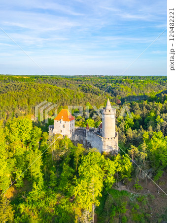 Kokorin Castle is bathed in golden sunlight as the evening approaches. Surrounded by lush green forests, the aerial view showcases its historic architecture and serene landscape. 129482221