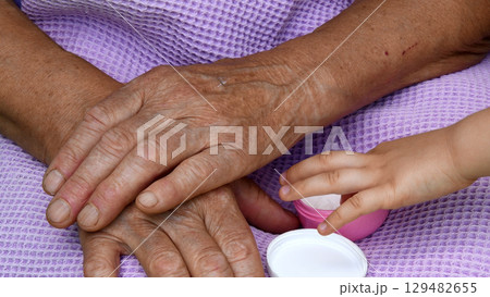 Child fingers take white cream from cosmetic jar. Wrinkled skin of granny hands closeup 129482655