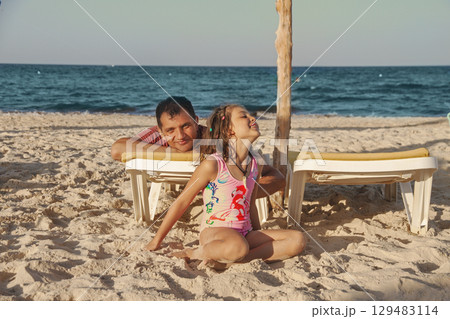 Dad and happy young daughter relaxing on sun lounger on beach by sea on hot day 129483114