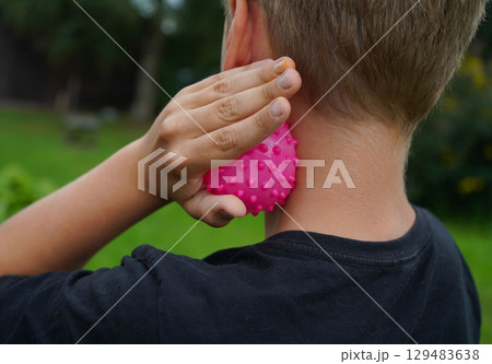 Photo from the back. The boy relaxes his neck muscles with a massage ball, running his hand over his neck. Photo from the back. The boy relaxes his neck muscles with a massage ball, running his hand over his neck. 129483638
