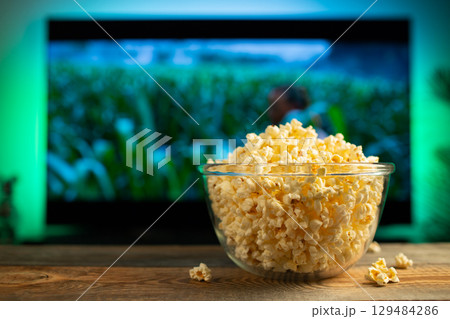 A glass bowl of popcorn and remote control in the background the TV works. Evening cozy watching a movie or TV series at home 129484286