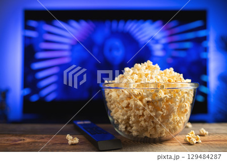 A glass bowl of popcorn and remote control in the background the TV works. Evening cozy watching a movie or TV series at home 129484287