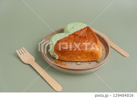 Heart shaped pistachio croissant on plate with recycled fork and knife on green background with copy space. Puff pastry trendy sweet dessert bakery. Creamy dough experiment  129484816