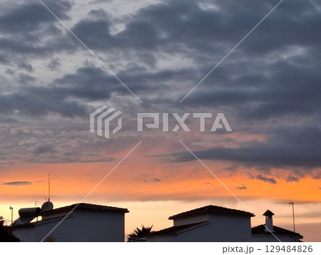 The sky is painted in vibrant shades of orange and purple as the sun sets behind tranquil rooftops. Clouds drift gently, creating a peaceful atmosphere in the neighborhood. 129484826