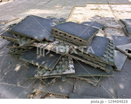 Workers are in the process of repairing the playground surface by removing old rubber tiles and stacking them. The setting is a community park under a bright sky, indicating mid-day. 129484828