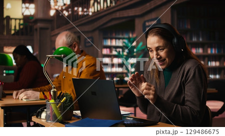 Woman listens to loud music on headset and disturbing other people in the library, senior man asking her to keep the volume low, breaking the library rules. Careless person singing. 129486675