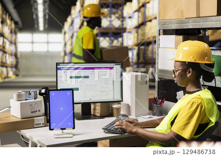 Black woman typing awb tracking info next to isolated mockup display, overseeing shipment details with hi vis vest and helmet in warehouse setting. Monitoring package tracking on software. Black woman typing awb tracking info next to isolated mockup display, overseeing shipment details with hi vis vest and helmet in warehouse setting. Monitoring package tracking on software. 129486738