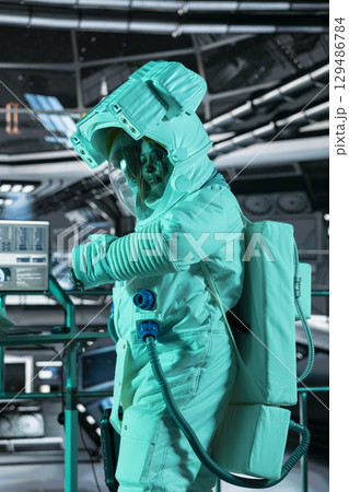 Female astronaut in spacesuit checks wrist device inside futuristic spacecraft while monitoring mission data on nearby display. Spacecraft crew member prepares for space travel operations 129486784