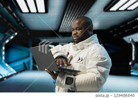 Astronaut wearing spacesuit inside futuristic spacecraft monitoring mission data on laptop screen. Spaceman wearing equipment prepares for space travel operations, reading data on notebook 129486840