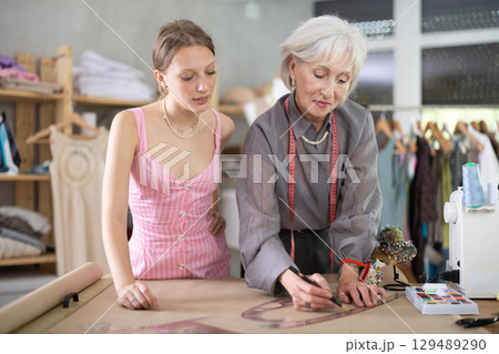 Positive mature tailoress showing young girl how to draw dress design 129489290