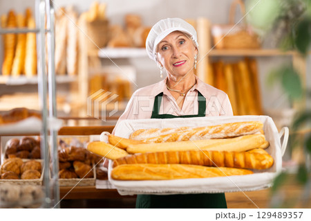 Senior female baker worker sells baguette, shows many products Senior female baker worker sells baguette, shows many products 129489357