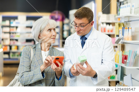 Old woman showing to middle-aged male pharmacist QR-code of product on her smartphone 129489547