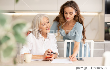 Positive woman and her senior mother sitting at table in kitchen, doing family financial paperwork Positive woman and her senior mother sitting at table in kitchen, doing family financial paperwork 129489729