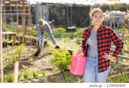 Happy young female holding pink watering can in hands while caring for vegetable and plant sprouts on sunny day in autumn Happy young female holding pink watering can in hands while caring for vegetable and plant sprouts on sunny day in autumn 129489905