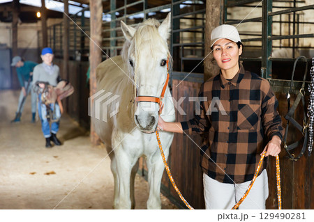 Female stable worker leads a white horse out of stall 129490281