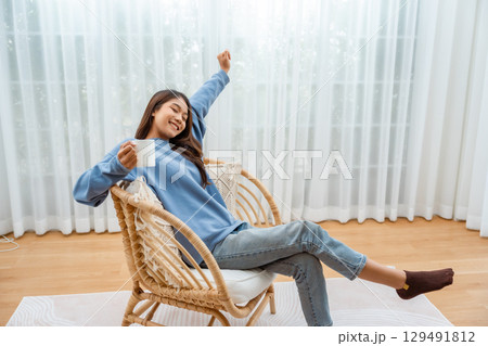 Young Asian woman happy and smiling, sitting on seat and holding mug in living room at home 129491812