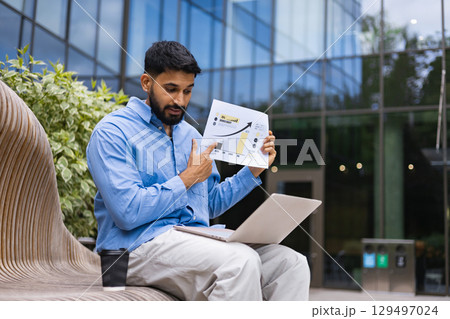 A businessman points to a chart during a presentation outdoors, using a laptop and coffee cup on a bench. A businessman points to a chart during a presentation outdoors, using a laptop and coffee cup on a bench. 129497024