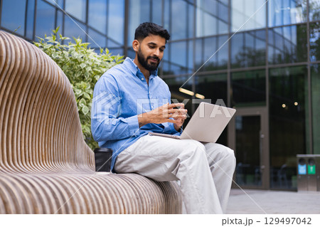 A man in a blue shirt sits on a wooden bench outside using a laptop, with a coffee cup beside him. 129497042