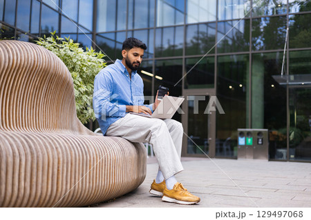 A man working on his laptop while sitting outside, holding a coffee cup. 129497068