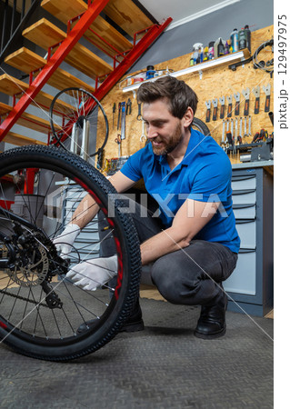 Bearded man servicing bike wheel in indoor workshop surrounded by tools drawers and industrial staircase during bicycle maintenance Bearded man servicing bike wheel in indoor workshop surrounded by tools drawers and industrial staircase during bicycle maintenance 129497975