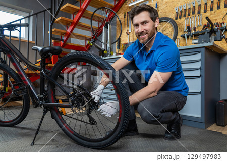 Bicycle mechanic in blue shirt kneeling with gloves adjusting cycle tire in organized indoor garage 129497983