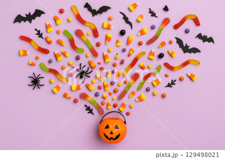 Halloween candy explosion with gummy worms, flying bats, spiders, and pumpkin bucket scattered soft violet background, creating festive atmosphere 129498021