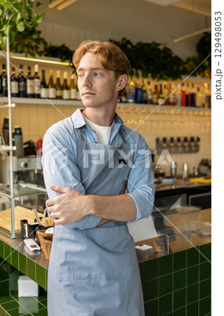 Pensive male barista in apron standing with crossed arms next to counter in stylish coffee shop Pensive male barista in apron standing with crossed arms next to counter in stylish coffee shop 129498053