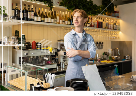 Cheerful man in apron posing near espresso bar standing with arms crossed in coffee shop interior 129498058
