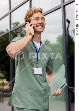 Caucasian male doctor in scrubs talking on smartphone near hospital Caucasian male doctor in scrubs talking on smartphone near hospital 129498081