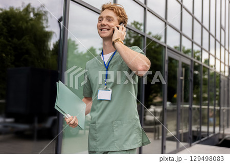 Ginger doctor standing by glass clinic building holding and discussing on smartphone Ginger doctor standing by glass clinic building holding and discussing on smartphone 129498083