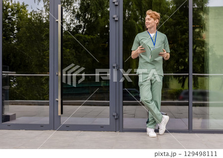 Red haired doctor in lab coat standing near glass clinic building holding medical folder 129498111