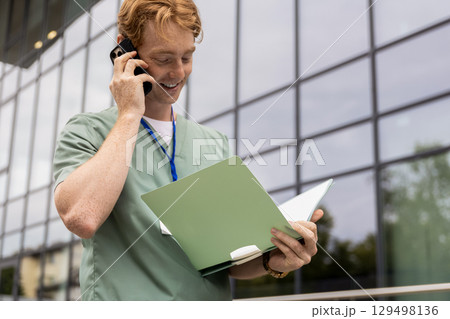 Ginger doctor standing by glass clinic building holding and discussing on smartphone Ginger doctor standing by glass clinic building holding and discussing on smartphone 129498136