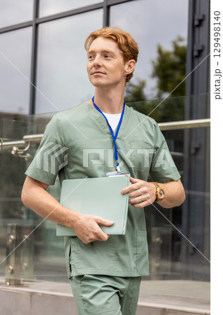 Red-haired healthcare worker in lab coat moving past glass building holding documents 129498140