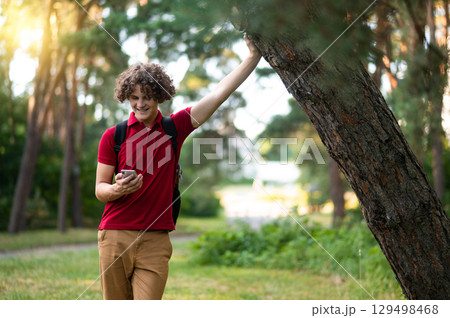 Curly-haired teen in red tshirt standing near the tree at the park 129498468