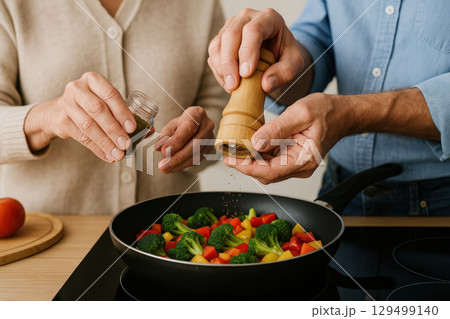 Senior couple cooking together adding spices to fresh vegetable stir fry in kitchen, warm and cozy atmosphere with healthy meal preparation 129499140