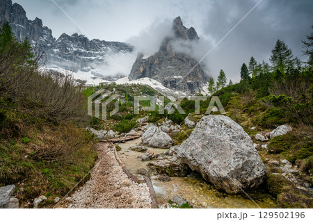 Rocky hiking trail near Lago Sorapis in the Dolomites (Dolomiti, Dolomiten), Italy. The path runs alongside a small mountain stream, with massive boulders and peaks in the background Rocky hiking trail near Lago Sorapis in the Dolomites (Dolomiti, Dolomiten), Italy. The path runs alongside a small mountain stream, with massive boulders and peaks in the background 129502196