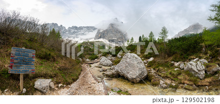 Rocky hiking trail near Lago Sorapis in the Dolomites (Dolomiti, Dolomiten), Italy. The path runs alongside a small mountain stream, with massive boulders and peaks in the background Rocky hiking trail near Lago Sorapis in the Dolomites (Dolomiti, Dolomiten), Italy. The path runs alongside a small mountain stream, with massive boulders and peaks in the background 129502198