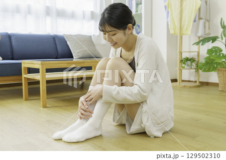 リビングのフローリングに座って靴下を足先を触る若い女性 129502310