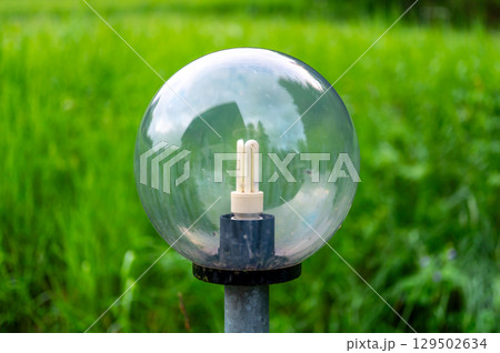 Outdoor light with large glass spheres housing fluorescent bulbs, illuminating the surroundings. In the background, a lush green meadow stretches under the clear sky 129502634
