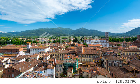 Panoramic view of Lucca, Italy, with red brick rooftops and historic churches seen from above. Tuscan hills and mountains form a stunning summer backdrop under a clear blue sky. Panoramic view of Lucca, Italy, with red brick rooftops and historic churches seen from above. Tuscan hills and mountains form a stunning summer backdrop under a clear blue sky. 129502790