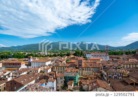 Panoramic view of Lucca, Italy, with red brick rooftops and historic churches seen from above. Tuscan hills and mountains form a stunning summer backdrop under a clear blue sky. 129502791