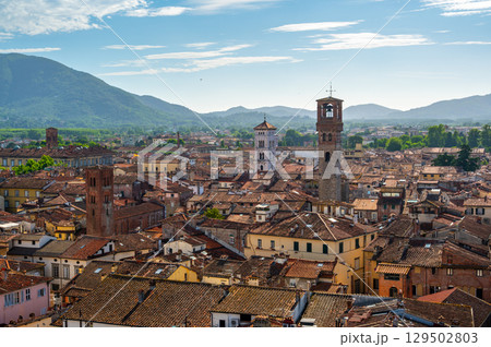 Panoramic view of Lucca, Italy, with red brick rooftops and historic churches seen from above. Tuscan hills and mountains form a stunning summer backdrop under a clear blue sky. Panoramic view of Lucca, Italy, with red brick rooftops and historic churches seen from above. Tuscan hills and mountains form a stunning summer backdrop under a clear blue sky. 129502803