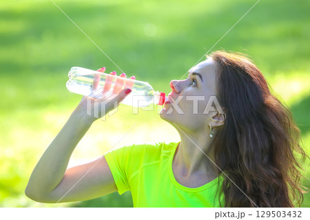 Woman drinking water while exercising outdoors in a vibrant green park setting 129503432
